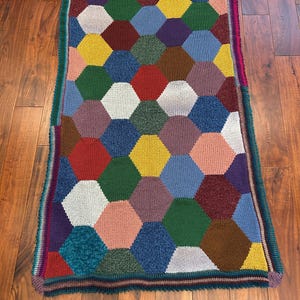 May include: A vibrant, handmade blanket with a patchwork design of hexagonal shapes in various colours, including red, yellow, green, and blue. The blanket has a dark green border with a striped edge. The blanket is made of a soft, knitted material.