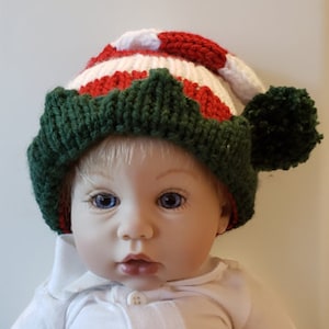 May include: A red, white, and green striped knitted elf hat with a white pom-pom on top. The hat has a green felt trim around the bottom.