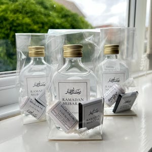 May include: Three clear glass bottles with gold lids and white labels that say "Ramadan Mubarak" in Arabic and English. Each bottle is in a clear plastic bag with a small white gift bag and a chocolate bar with the same label.