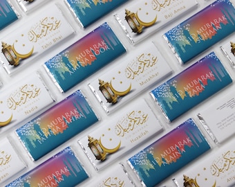 Puede incluir: Surtido de envoltorios de barras de chocolate de colores con caligrafía árabe y el texto "Eid Mubarak" seguido de un nombre. Los envoltorios presentan una luna creciente y un diseño de linterna.