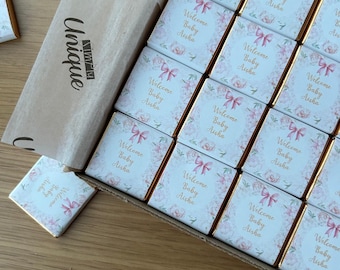 May include: A box of small, white chocolate bars with pink floral designs and gold trim. Each bar has the text "Welcome Baby Aisha" printed on it.