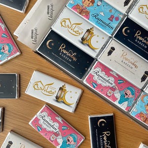 Op de afbeelding: Een assortiment chocolade repen verpakt in folie met verschillende Ramadan Kareem-ontwerpen. De ontwerpen bevatten illustraties van lantaarns, sterren en halve manen, samen met Arabische kalligrafie. De repen zijn gerangschikt op een houten oppervlak.