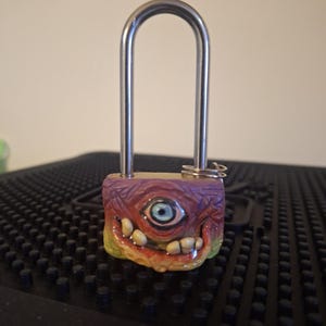 May include: A colorful, ceramic padlock with a single eye and teeth. The padlock is attached to a silver metal loop.
