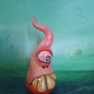 May include: A whimsical, hand-painted sculpture of a creature with a pink body, a large eye, and a yellow, tooth-like structure. The sculpture has a curved, horn-like extension and is set against a green background.