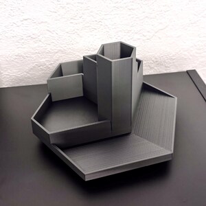 Könnte beinhalten: Ein grauer sechseckiger Schreibtisch-Organizer mit mehreren Fächern. Der Organizer hat ein hohes zentrales Fach und zwei kleinere Fächer auf jeder Seite. Der Organizer ist aus Kunststoff und hat eine glatte, matte Oberfläche.