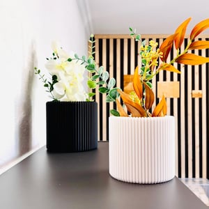 Può includere: Due fioriere decorative, una nera e una bianca, ciascuna riempita con fiori e verde artificiali. Le fioriere hanno un design a coste. La fioriera nera contiene fiori bianchi e foglie verdi, mentre la fioriera bianca ha un fogliame arancione e giallo.