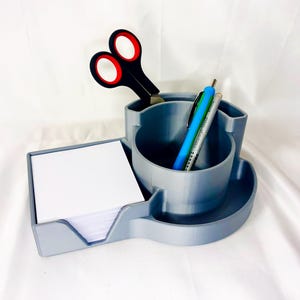 Può includere: Un organizzatore da scrivania grigio con un vano per penne e matite, un vano per appunti adesivi e un bicchiere per forbici. L'organizzatore è in plastica e ha un design moderno.