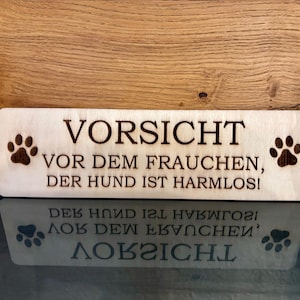 Könnte beinhalten: Ein Holzschild mit dem Text "VORSICHT VOR DEM FRAUCHEN, DER HUND IST HARMLOS!" in schwarzer Schrift. Das Schild hat zwei Pfotenabdrücke auf jeder Seite des Textes.