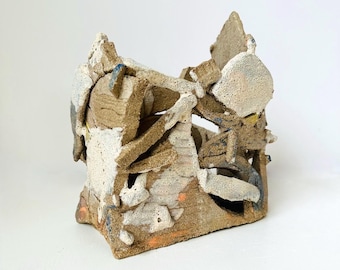 Puede incluir: Una pequeña escultura abstracta de cerámica con una superficie rugosa y texturizada. La escultura está hecha de arcilla marrón, blanca y naranja y tiene una forma geométrica.