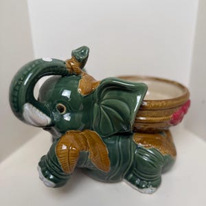 May include: A ceramic elephant figurine in a dark green glaze with brown accents on the ears, trunk, and back. The elephant features a raised trunk and a circular, basket-weave patterned bowl attached to its back, decorated with small red floral details.