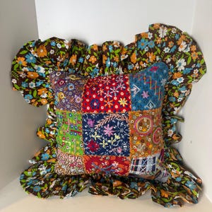 May include: A square throw pillow featuring a patchwork center of various embroidered fabric squares in red, blue, green, and brown. The pillow is bordered by a wide, ruffled trim made of brown fabric with a repeating floral print in blue, orange, and white.