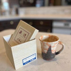 May include: A handmade ceramic mug with a handle, featuring a blend of orange, black, and white colors. Beside it is a wooden box containing tea, with Japanese text on the box and a blue label.