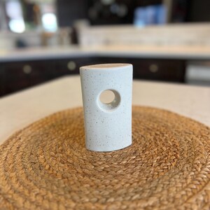 May include: White ceramic candle holder with a speckled finish and a circular cutout. The holder is sitting on a woven brown mat.