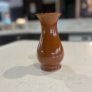 May include: A brown ceramic vase with a glossy finish. The vase has a rounded body, a flared top, and a small base. The vase is sitting on a white countertop. The vase is likely used for displaying flowers or other decorative items.