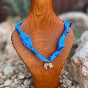 May include: A blue ribbon necklace with a gold-colored angel wing pendant. The necklace is displayed on a wooden bust. The background features rocks and greenery.