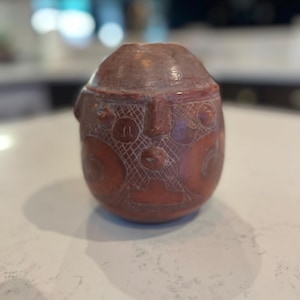 May include: A reddish-brown, egg-shaped sculpture with carved facial features and geometric patterns. The sculpture has a rounded top and a detailed face with etched lines and circles. The object is likely a decorative piece or a historical artifact.
