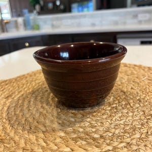 May include: A dark brown ceramic bowl with a smooth, glossy finish. The bowl has a rounded shape and a slightly flared rim. It is sitting on a woven straw placemat.