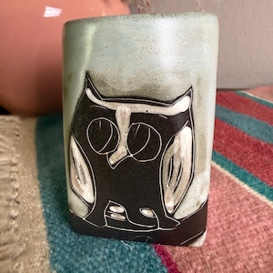 May include: A cylindrical ceramic vase featuring a stylized black owl graphic with white line details set against a mottled light blue and grey background. The surface has a smooth finish with visible texture and subtle color variations throughout the piece.