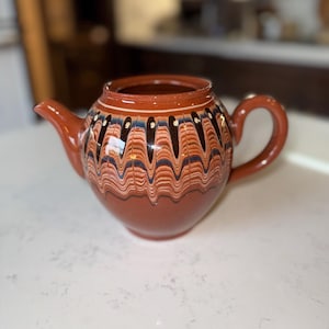 May include: A ceramic teapot with a brown base and a decorative band of blue, black, and white wavy lines. The teapot has a curved handle and a spout. The background is blurred.