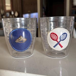 May include: Two clear double-walled tumblers. One tumbler features a blue patch with a gold horse head and horseshoe design. The other has a white patch with crossed red and blue tennis rackets. The tumblers are sitting on a white surface.