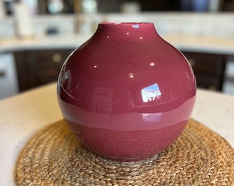 May include: A burgundy-colored, round vase with a textured lower half and a smooth, glossy upper half. The vase sits on a round, woven placemat. The vase has a wide opening and a bulbous shape, suitable for home decor.