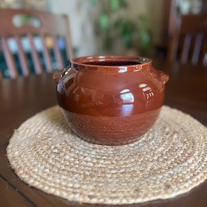 May include: A brown ceramic pot with two handles. The pot is sitting on a woven straw placemat.