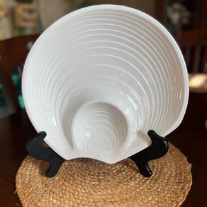 May include: White ceramic bowl with a unique, layered design. The bowl is on a black stand and a woven mat.
