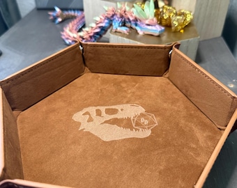 May include: A brown leather dice tray with a dinosaur skull and a D20 die graphic in the center. The tray has a hexagonal shape with raised sides and metal snap closures. Other fantasy figurines are in the background.