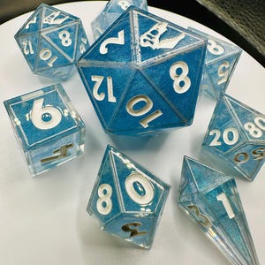 May include: A collection of blue and clear resin dice with white numbering. The dice are various shapes, including a 20-sided die, a 12-sided die, and a 6-sided die. The dice have a glittery, iridescent effect.