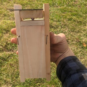 Op de afbeelding: Een lichtgekleurd houten apparaat met een rechthoekig lichaam en een scharnierend bovendeel. Het apparaat heeft metalen haken en een klein, donkergekleurd mes. Het apparaat wordt tegen een achtergrond van groen gras gehouden.