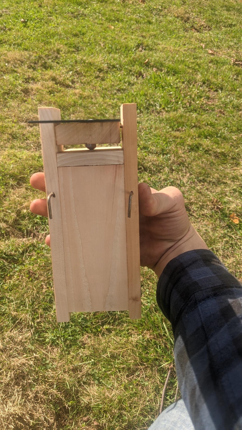 Op de afbeelding: Een lichtgekleurd houten apparaat met een rechthoekig lichaam en een scharnierend bovendeel. Het apparaat heeft metalen haken en een klein, donkergekleurd mes. Het apparaat wordt tegen een achtergrond van groen gras gehouden.