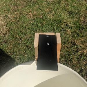 Op de afbeelding: Een houten constructie met een zwart rechthoekig stuk bevestigd, gepositioneerd boven een witte, afgeronde container. De constructie is bevestigd met twee zilveren bevestigingsmiddelen. De sc&egrave;ne speelt zich buiten af op een grasachtig oppervlak.