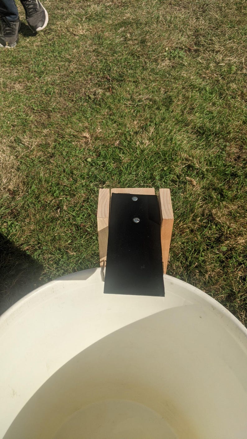 Op de afbeelding: Een houten constructie met een zwart rechthoekig stuk bevestigd, gepositioneerd boven een witte, afgeronde container. De constructie is bevestigd met twee zilveren bevestigingsmiddelen. De sc&egrave;ne speelt zich buiten af op een grasachtig oppervlak.