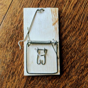 May include: A wooden mousetrap with a metal spring and wire components. The trap is rectangular and made of light-colored wood, with a metal spring mechanism designed to snap shut. The trap is set on a wooden surface.