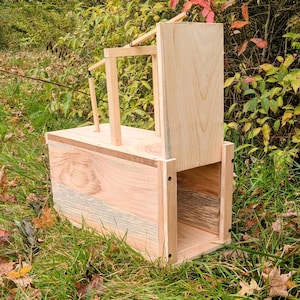 May include: A wooden birdhouse with a rectangular base and a sloped roof. The birdhouse is made of light-colored wood and has a natural finish. The birdhouse is sitting on a bed of green grass.