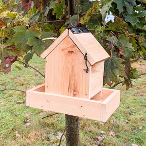Könnte beinhalten: Ein Vogelhaus aus Holz mit Satteldach und Futtertablett. Das Vogelhaus ist aus hellfarbenem Holz gefertigt und hat einen schwarzen Metallverschluss. Es hängt an einem Ast mit grünen und gelben Blättern.