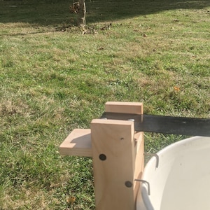 Op de afbeelding: Een houten apparaat met een metalen mes en een witte emmer. Het apparaat is met metalen haken aan de emmer bevestigd. Het apparaat staat op een grasveld met bomen op de achtergrond.