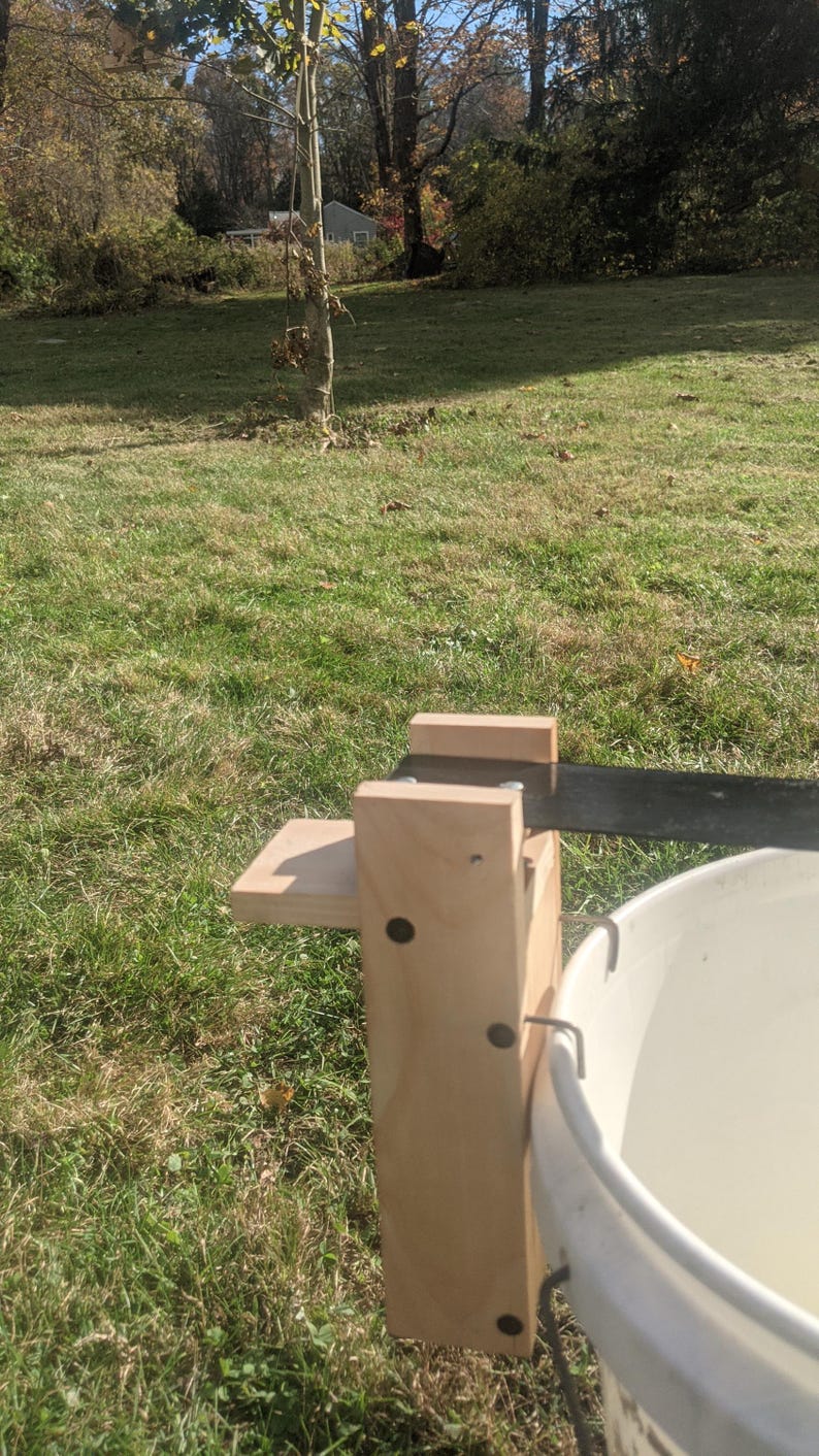 Op de afbeelding: Een houten apparaat met een metalen mes en een witte emmer. Het apparaat is met metalen haken aan de emmer bevestigd. Het apparaat staat op een grasveld met bomen op de achtergrond.