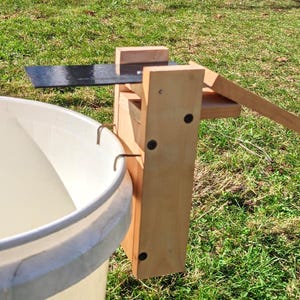 Op de afbeelding: Een houten emmerval met een metalen mes en een witte emmer. De val is gemaakt van lichtgekleurd hout en is ontworpen om op de rand van de emmer te zitten. Het metalen mes is zwart en steekt boven de emmer uit.