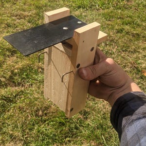 Op de afbeelding: Een houten vogelhuisje met een zwart metalen dak en twee metalen haken. Het vogelhuisje is gemaakt van lichtgekleurd hout en wordt door een persoon vastgehouden tegen een achtergrond van groen gras.