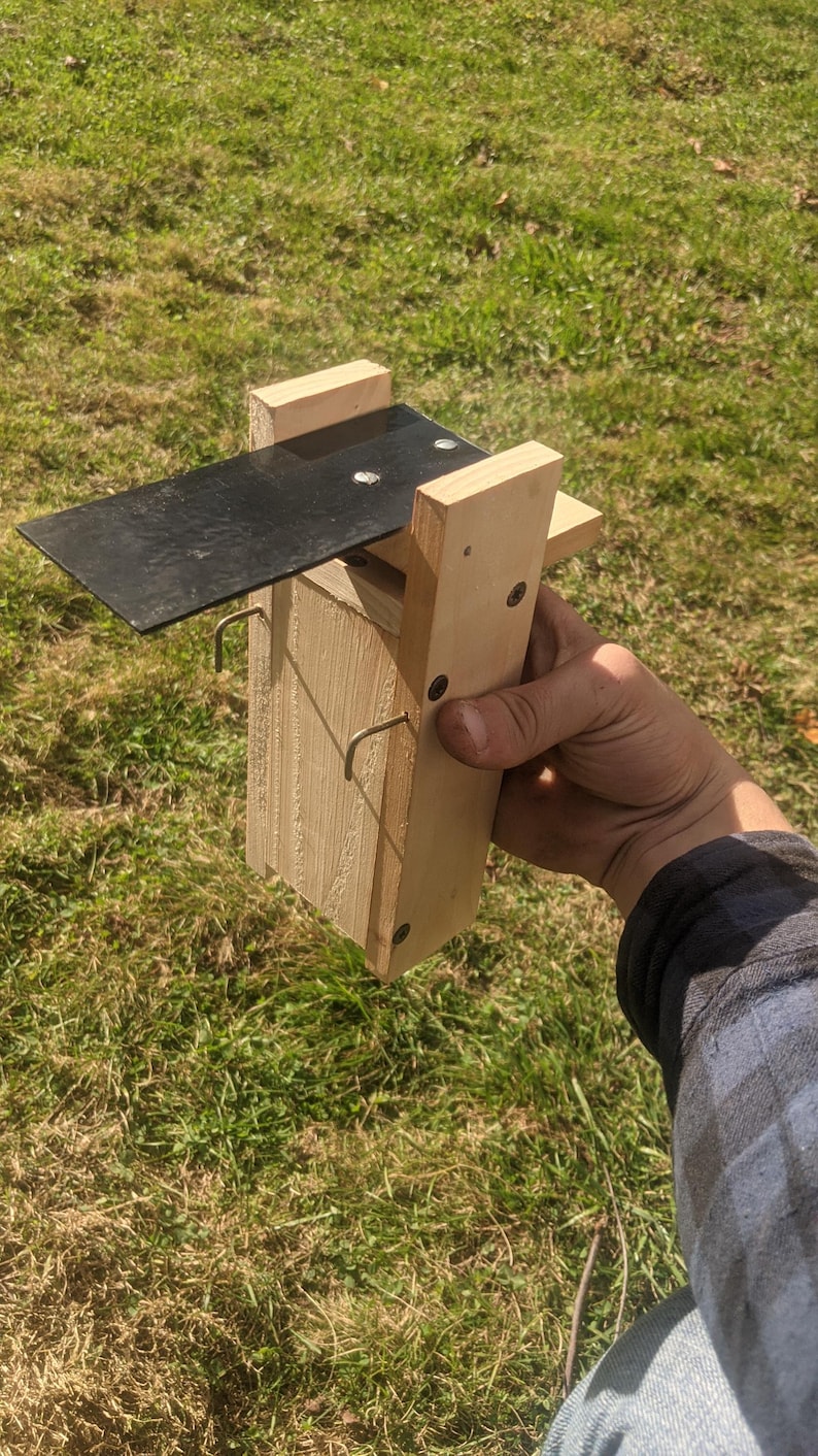 Op de afbeelding: Een houten vogelhuisje met een zwart metalen dak en twee metalen haken. Het vogelhuisje is gemaakt van lichtgekleurd hout en wordt door een persoon vastgehouden tegen een achtergrond van groen gras.