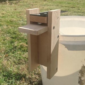 Op de afbeelding: Een houten emmerdekselheffer, bevestigd aan een witte emmer. De heffer is gemaakt van lichtgekleurd hout en heeft een klein platform en een metalen scharnier. Het apparaat is ontworpen om een emmerdeksel op te tillen.