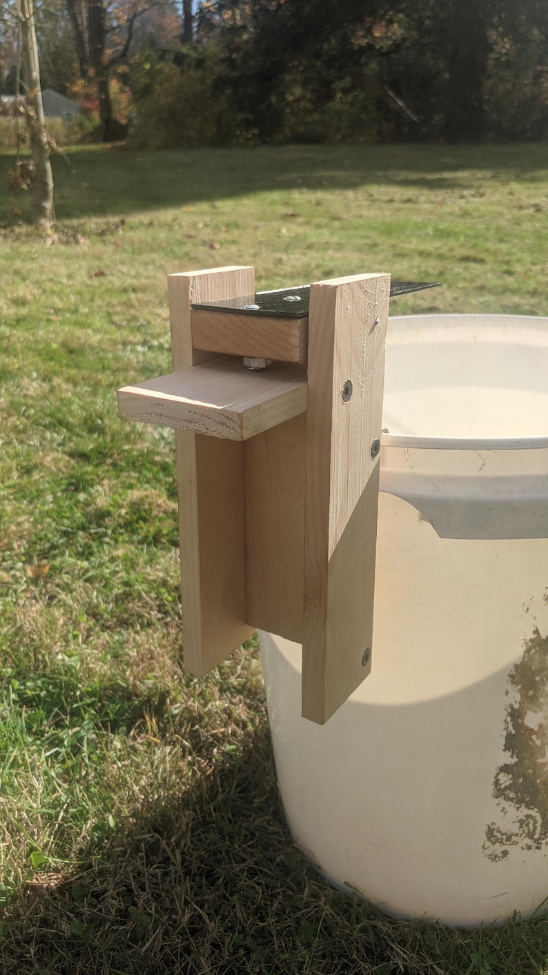 Op de afbeelding: Een houten emmerdekselheffer, bevestigd aan een witte emmer. De heffer is gemaakt van lichtgekleurd hout en heeft een klein platform en een metalen scharnier. Het apparaat is ontworpen om een emmerdeksel op te tillen.