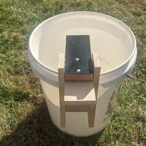 Op de afbeelding: Een zelfgemaakte houten helling en platform, met een zwarte bovenkant, is in een witte emmer geplaatst. De constructie is gemaakt van lichtgekleurd hout en is met schroeven bevestigd. De emmer staat op groen gras.