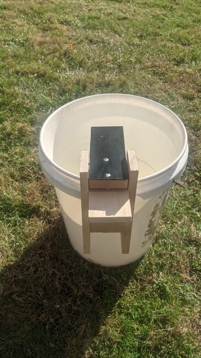 Op de afbeelding: Een zelfgemaakte houten helling en platform, met een zwarte bovenkant, is in een witte emmer geplaatst. De constructie is gemaakt van lichtgekleurd hout en is met schroeven bevestigd. De emmer staat op groen gras.
