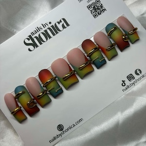 May include: A set of ten artificial nails with a gradient design in shades of red, orange, yellow, green, and blue. Each nail features a gold-colored ring accent. The nails are displayed on a white card with the text "nails by Shonica".