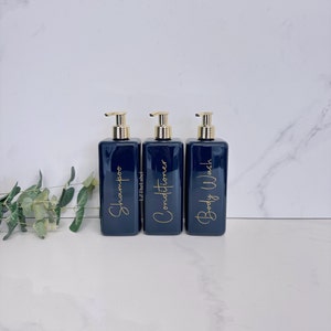 K&ouml;nnte beinhalten: Drei dunkelblaue Pumpflaschen mit goldenen Verschl&uuml;ssen und Etiketten. Die Etiketten zeigen "Shampoo", "Conditioner" und "Duschgel" in goldener Schrift.