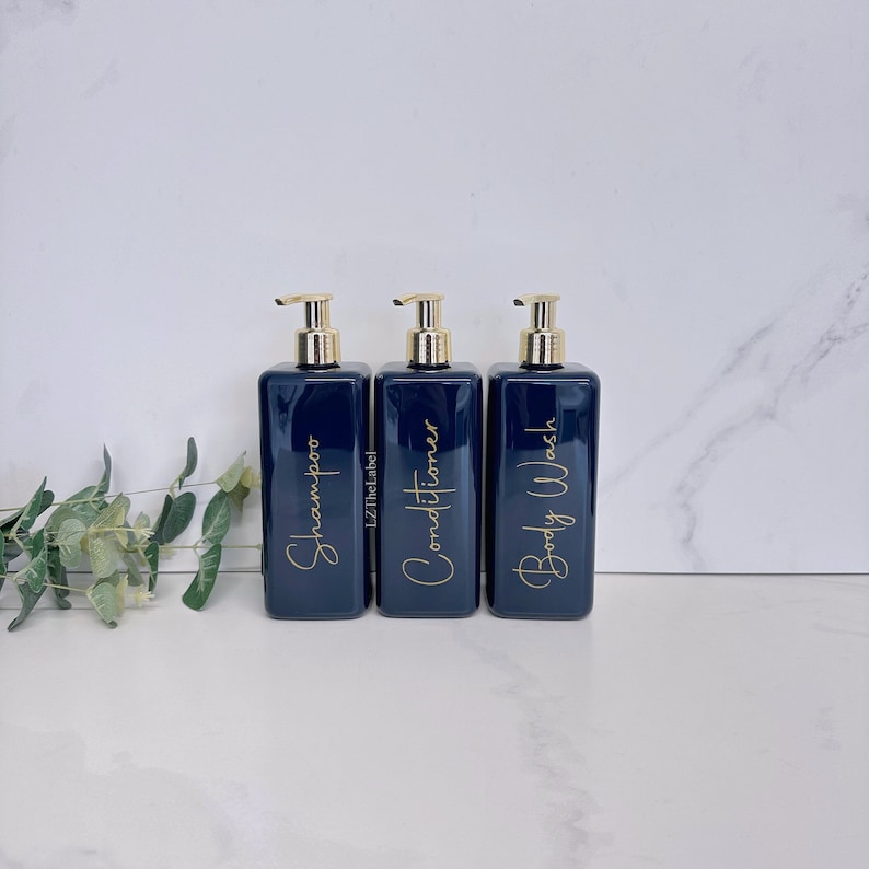 K&ouml;nnte beinhalten: Drei dunkelblaue Pumpflaschen mit goldenen Verschl&uuml;ssen und Etiketten. Die Etiketten zeigen "Shampoo", "Conditioner" und "Duschgel" in goldener Schrift.