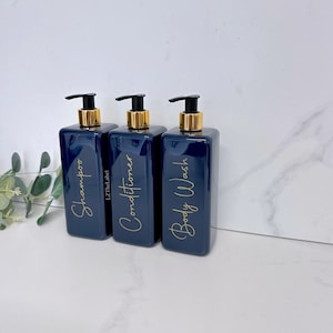 K&ouml;nnte beinhalten: Drei marineblaue Pumpflaschen mit goldenen Deckeln und Etiketten. Die Etiketten sind in goldener Schrift geschrieben und sagen "Shampoo", "Conditioner" und "Duschgel".