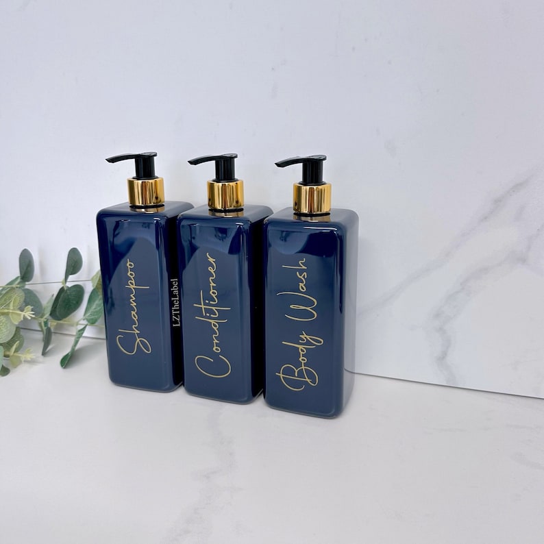 K&ouml;nnte beinhalten: Drei marineblaue Pumpflaschen mit goldenen Deckeln und Etiketten. Die Etiketten sind in goldener Schrift geschrieben und sagen "Shampoo", "Conditioner" und "Duschgel".